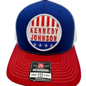 May include: A blue and red trucker hat with a white mesh back. The front features a circular patch with red vertical stripes, the text KENNEDY JOHNSON in red, and four white stars on a blue background. A black sticker on the brim reads RICHARDSON 112.