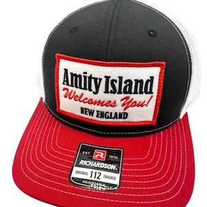 Puede incluir: Una gorra tipo camionero negra y blanca con visera roja y un parche rectangular al frente. El parche tiene el texto Amity Island Welcomes You! NEW ENGLAND en letras negras y rojas. Una etiqueta negra en la visera dice RICHARDSON 112 TRUCKER.