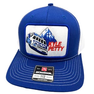 May include: A royal blue and white trucker hat featuring a rectangular patch on the front. The patch displays a white race car with the text PEAK, a blue mountain graphic, and the red text KYLE PETTY. A black sticker on the brim reads RICHARDSON 112.