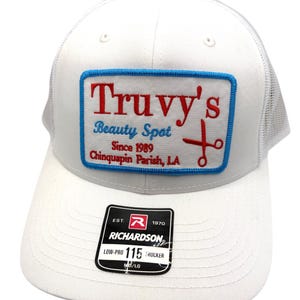 May include: A white trucker hat featuring a rectangular patch with a blue border. The patch displays red and blue text reading Truvy's Beauty Spot Since 1989 Chinquapin Parish, LA, with a red scissors icon. A black Richardson 115 sticker is on the brim.