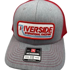 Peut inclure: Une casquette trucker grise et rouge avec un écusson rectangulaire portant le texte Riverside International Raceway en rouge et bleu. La casquette présente un panneau avant gris, un filet arrière rouge et une visière grise avec des coutures rouges.