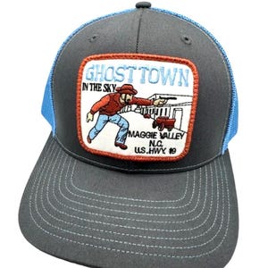 Puede incluir: Una gorra de béisbol gris carbón con una parte trasera de malla azul brillante y un parche rectangular en la parte delantera. El parche presenta una ilustración bordada de una persona con sombrero de vaquero y camisa roja con el texto GHOST TOWN IN THE SKY MAGGIE VALLEY N.C. U.S.HWY 19.