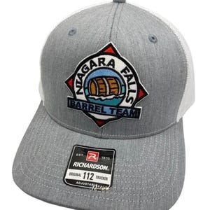 May include: A grey and white trucker hat featuring an embroidered patch on the front with the text Niagara Falls Barrel Team and an illustration of a wooden barrel. A black sticker on the brim displays the Richardson 112 Original logo.