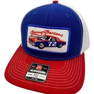 May include: A blue and white trucker hat with a red brim and a red button on top. The front features a rectangular patch with a red, white, and blue race car, the number 72, and the text Benny Parsons. A black sticker reads Richardson 112 Trucker.