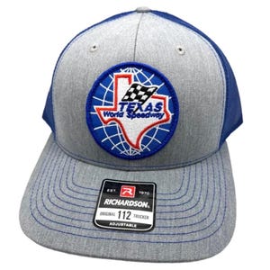 Könnte beinhalten: Eine graumelierte und blaue Trucker-Cap mit einem runden Aufnäher, der eine weiße Karte von Texas, eine Zielflagge und den Text Texas World Speedway zeigt. Ein schwarzer Aufkleber auf dem Schirm zeigt das Richardson 112 Trucker Logo.