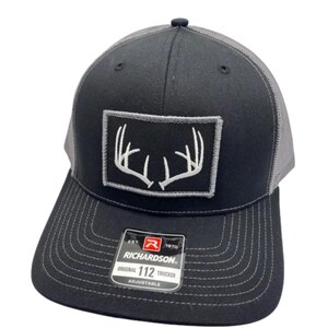 May include: A black and grey trucker hat featuring a rectangular patch on the front with a white embroidered deer antler design. The brim has a sticker that reads EST. 1970 RICHARDSON ORIGINAL 112 TRUCKER ADJUSTABLE. The back panels are grey mesh.