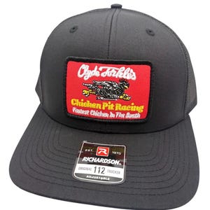 Puede incluir: Una gorra de camionero negra con un parche rectangular de fondo rojo. El parche muestra un gráfico de un pollo blanco y el texto "Clyde Forkle's", "Chicken Pit Racing" y "Fastest Chicken In The South". Una pegatina Richardson 112 está en la visera.