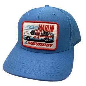May include: A light blue trucker hat featuring a rectangular patch with a red border. The patch displays a white race car with the number 44, the text Sterling Marlin, and the word Piedmont in red lettering against a white background.