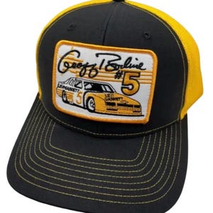 May include: A black and yellow trucker cap featuring a rectangular patch on the front. The patch displays a yellow race car with the number 5, the text Levi Garrett, and the signature Geoff Bodine #5 in black script against a white background.