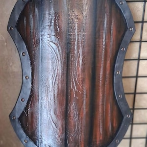 Könnte beinhalten: Ein Holzschild mit dunkelbraunem Finish und Metallrand. Der Schild hat eine gebogene Form mit einem strukturierten Holzmaserungsmuster. Der Metallrand ist dunkelgrau und mit Nieten befestigt.