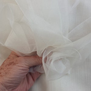 Puede incluir: Un primer plano de una tela blanca y transparente. La tela es suave y delicada, con una textura ligeramente arrugada. Se sostiene en una mano, y la tela se drapea sobre sí misma, creando una forma suave y fluida.