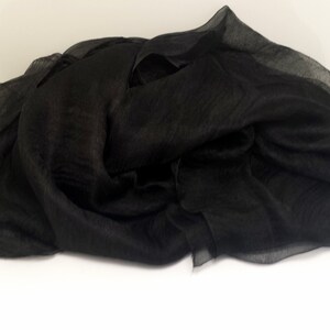 Puede incluir: Una bufanda negra drapeada, hecha de una tela ligera y transparente. La bufanda est&aacute; doblada y colocada sobre una superficie blanca, mostrando su textura fluida y su color oscuro. La bufanda parece ser de seda o un material similar.