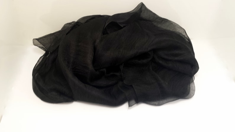 Puede incluir: Una bufanda negra drapeada, hecha de una tela ligera y transparente. La bufanda est&aacute; doblada y colocada sobre una superficie blanca, mostrando su textura fluida y su color oscuro. La bufanda parece ser de seda o un material similar.