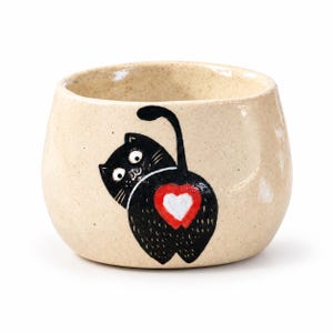 May include: A beige speckled ceramic bowl featuring a hand-painted illustration of a black cat looking backward. The cat has wide eyes and a long tail, with a red and white heart shape positioned on its rear. The bowl has a rounded shape.