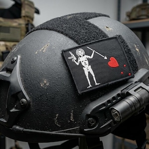 May include: A matte black tactical helmet featuring a rectangular embroidered patch. The patch displays a white skeleton figure holding a spear pointed toward a red heart with three red drops. A black tactical flashlight is mounted on the side rail.