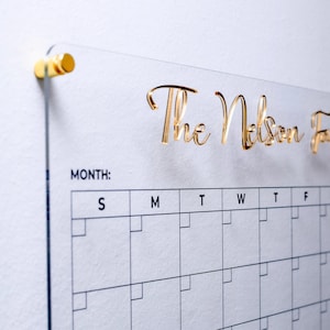 May include: A clear acrylic wall calendar featuring the gold text The Nelson Family at the top. The calendar includes a grid with the letters S, M, T, W, T, F, S for days of the week, a section labelled MONTH, and a side column for To Do lists.