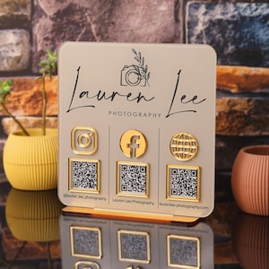 Puede incluir: Un letrero cuadrado de acrílico beige con el texto negro Lauren Lee Photography y un icono de cámara. Incluye tres iconos dorados de redes sociales y tres códigos QR correspondientes con marcos dorados sobre una base de soporte dorada.