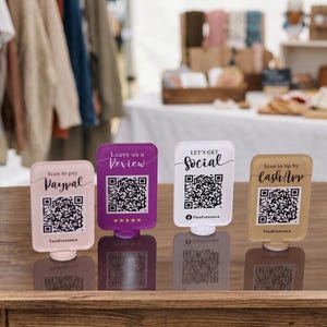 Peut inclure: Quatre panneaux rectangulaires en acrylique avec des codes QR posés sur une surface en bois. Les panneaux sont rose clair, violets, blancs et beiges, avec des textes comme Scan to pay Paypal, Leave us a Review, LET'S GET Social et Scan to tip by CashApp.
