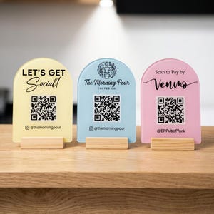 Puede incluir: Tres letreros acrílicos arqueados en amarillo pastel, azul claro y rosa suave, montados sobre bases de madera rectangulares. Los letreros tienen texto y códigos QR impresos en negro. El amarillo dice LET'S GET Social! y @themorningpour. El azul muestra un logo de café, The Morning Pour, y @themorningpour. El rosa dice Scan to Pay by Venmo y @EPPubofYork.