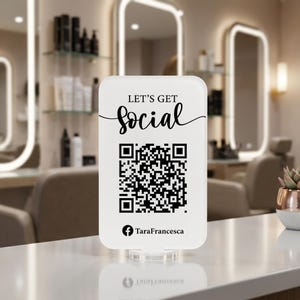 Peut inclure: Un panneau rectangulaire en acrylique blanc aux coins arrondis, présentant le texte noir LET'S GET SOCIAL au-dessus d'un code QR central et de l'icône Facebook avec le texte TaraFrancesca en bas, monté sur une base circulaire transparente.