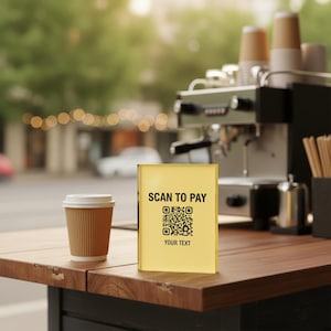 Puede incluir: Un letrero rectangular de acrílico amarillo con el texto negro SCAN TO PAY, un código QR central y las palabras YOUR TEXT en la parte inferior. El letrero se encuentra sobre una superficie de madera junto a un vaso de café de papel marrón.