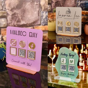 Puede incluir: Tres letreros de acrílico de formas cuadradas y orgánicas con códigos QR para redes sociales y pagos. Los letreros son de color morado claro, beige y verde claro, cada uno con iconos dorados y texto impreso para branding y pagos.
