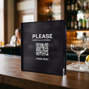 Puede incluir: Un letrero cuadrado de acrílico negro con texto blanco que dice PLEASE Leave us a review, un código QR central y las palabras YOUR TEXT en la parte inferior. El letrero tiene una superficie lisa y reflectante diseñada para superficies planas.