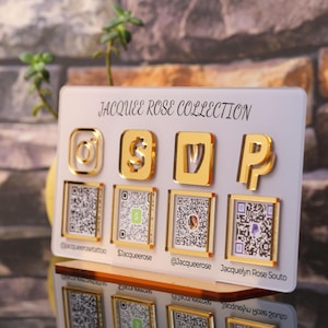 Custom acrylic QR code sign with four QR codes, gold social media icons and personalized business branding, displayed on a reflective countertop