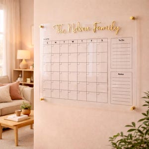 May include: A clear acrylic wall calendar featuring a grid for the month, a section for To Do, and a section for Notes. The text The Nelson Family is displayed in gold script at the top. The board is mounted with four gold-coloured metal standoffs.