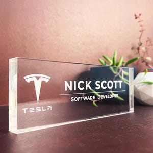 May include: A rectangular clear acrylic desk nameplate featuring the white Tesla logo and the text "NICK SCOTT" above the title "SOFTWARE DEVELOPER" in white lettering. The nameplate has a thick, transparent block design with polished edges.
