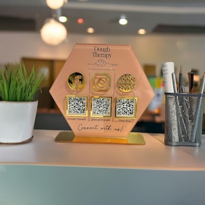 Custom acrylic QR code sign with three QR codes, gold social media icons and personalized business branding, displayed on a reflective countertop