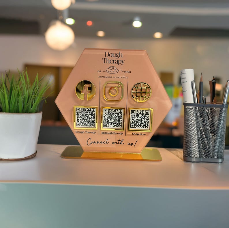 Custom acrylic QR code sign with three QR codes, gold social media icons and personalized business branding, displayed on a reflective countertop