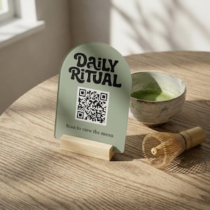 Op de afbeelding: Een boogvormig saliegroen acrylbord met een houten voet, voorzien van de zwarte tekst Daily Ritual, een QR-code en de tekst Scan to view the menu. Het bord staat naast een keramische kom met groene thee en een bamboe garde.