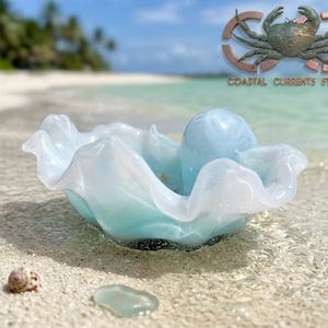 May include: A light blue and white glass bowl with a ruffled, wavy edge and a smooth, rounded interior protrusion. The glass features a swirling, marbled color pattern that transitions from a pale blue base to a translucent white rim.