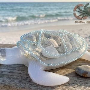 May include: A translucent white sea turtle figurine with a shell filled with various small white seashells. A delicate net of light blue beads is draped over the turtle's shell. The turtle is positioned on a weathered wooden surface.