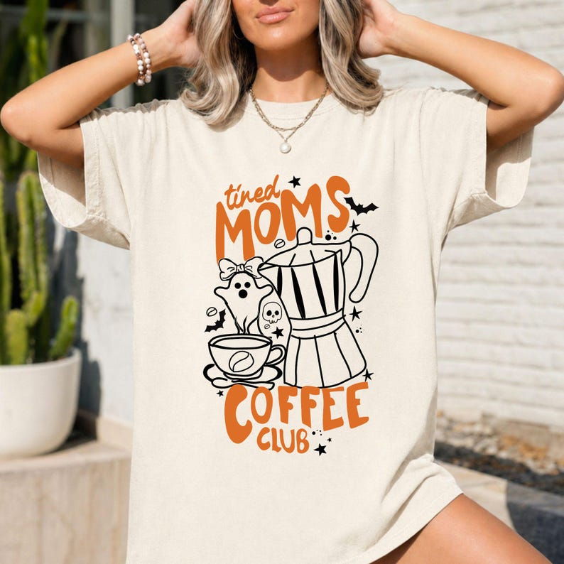 A woman wearing a white t-shirt with a graphic design featuring a coffee maker, a mug, and various Halloween-themed elements.