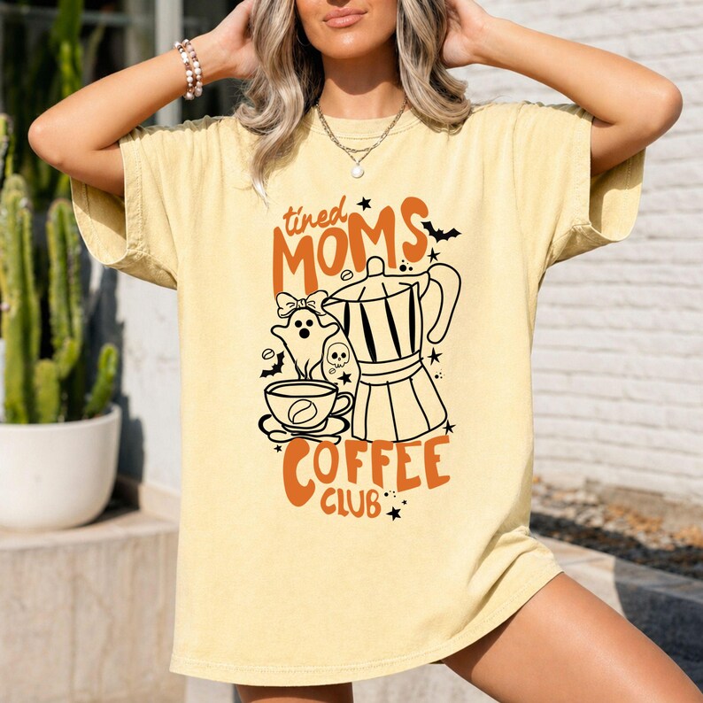 A woman wearing a yellow t-shirt with a graphic design featuring a coffee pot, a ghost, and the text "tired Moms Coffee Club".