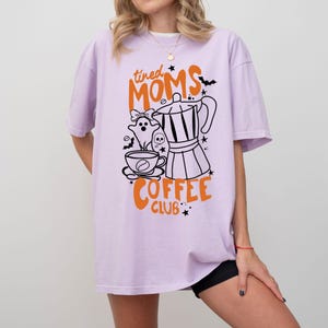 A woman wearing a purple t-shirt with a graphic design featuring a coffee pot, ghost, and the text "tired Moms Coffee Club" printed on it.
