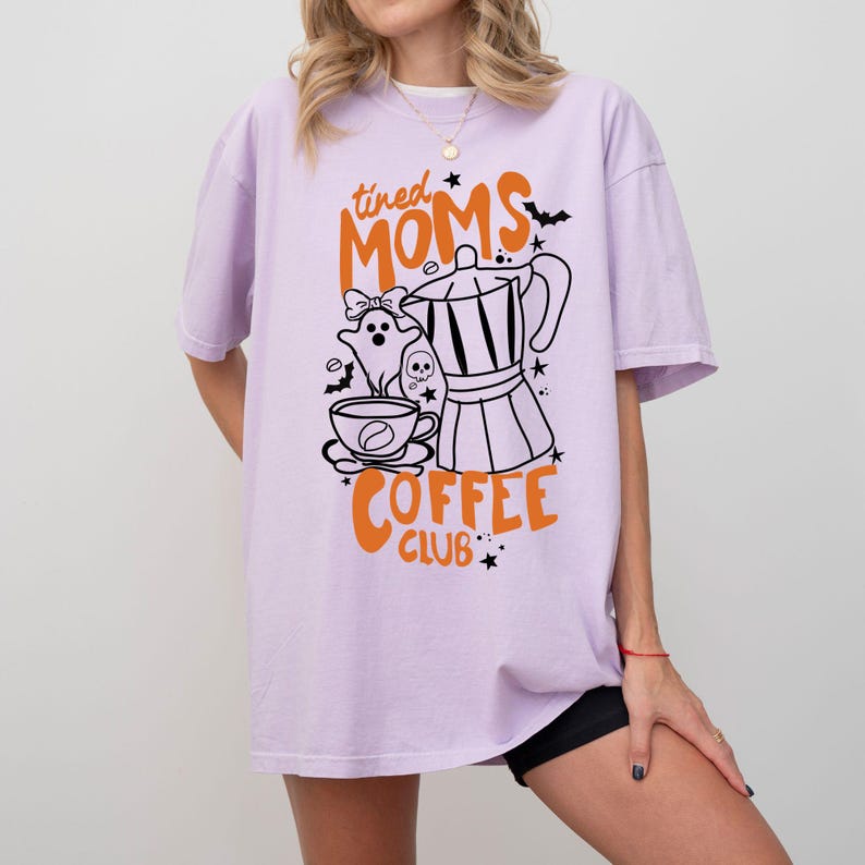 A woman wearing a purple t-shirt with a graphic design featuring a coffee pot, ghost, and the text "tired Moms Coffee Club" printed on it.