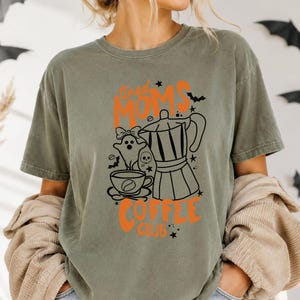 A woman wearing a green t-shirt with a graphic design featuring a coffee pot, a ghost, and the text "Tired Moms Coffee Club" printed on it.