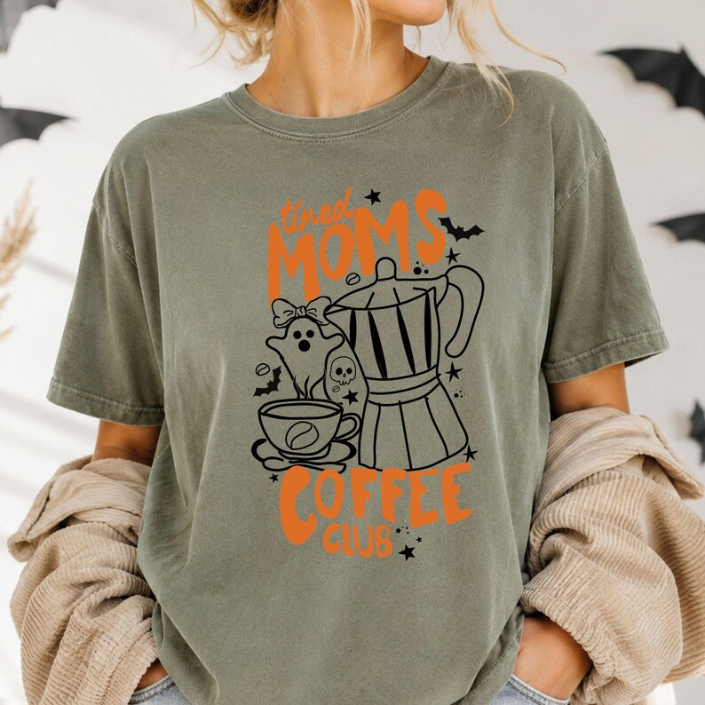 A woman wearing a green t-shirt with a graphic design featuring a coffee pot, a ghost, and the text "Tired Moms Coffee Club" printed on it.