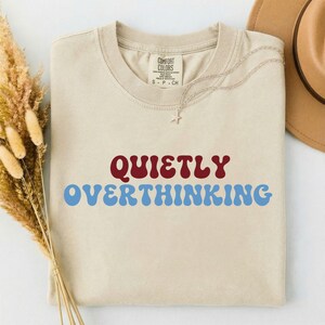May include: A beige short-sleeved t-shirt with a crew neckline featuring the text QUIETLY OVERTHINKING in dark red and light blue wavy lettering. The shirt has a Comfort Colors label at the inner neck and a silver star-shaped pendant necklace.