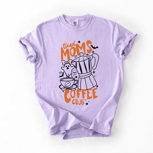 A purple t-shirt with a graphic design featuring a coffee pot, ghost, and the text "tired Moms Coffee Club" in orange and black.