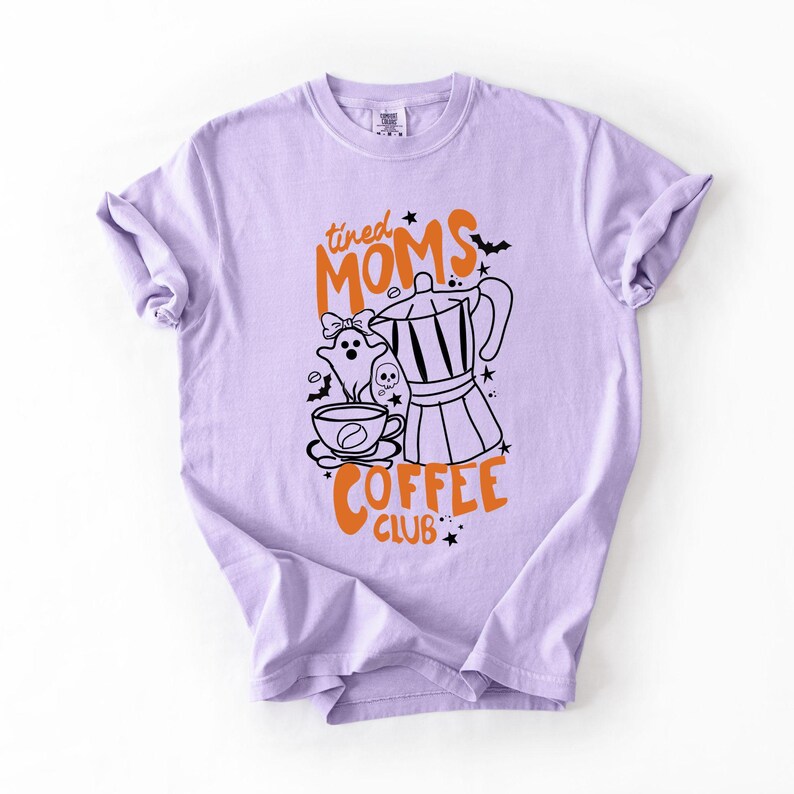 A purple t-shirt with a graphic design featuring a coffee pot, ghost, and the text "tired Moms Coffee Club" in orange and black.