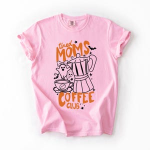 A pink t-shirt with a graphic design featuring a coffee maker, a ghost, and the text "tired Moms Coffee Club" printed on it.