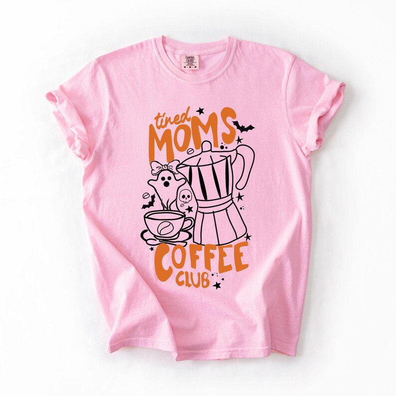 A pink t-shirt with a graphic design featuring a coffee maker, a ghost, and the text "tired Moms Coffee Club" printed on it.