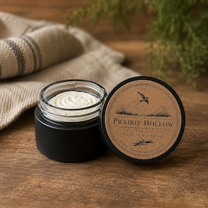 May include: A black jar containing white cream with a circular label that reads Prairie Hollow Handcrafted Goat Milk Soap Vanilla Cream Rooted in Nature Made to Nurture, featuring an illustration of a bird flying over tall grass.