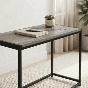 May include: A rectangular desk featuring a gray wood-grain surface and a black metal frame. A closed notebook with a black pen resting on top and a small potted succulent plant are placed on the desk surface, along with a pair of thin-rimmed glasses.