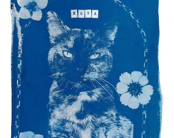 May include: A blue and white cyanotype print on paper featuring a portrait of a cat centred within an arched frame. The word NOVA is printed in white block letters above the cat, surrounded by three white floral illustrations on the blue background.