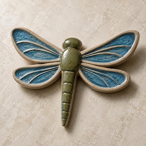 May include: A ceramic dragonfly wall ornament featuring a glossy olive green body and four textured blue wings with beige outlines. The insect-shaped piece has a segmented tail and a smooth, rounded head, showcasing a handcrafted glazed finish.